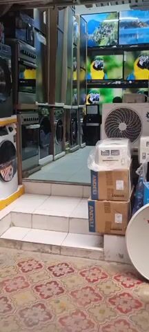The video opens with a view of an electronics store, displaying various appliances like washing machines and televisions. A man walks across the screen from left to right. The store is filled with products, including boxes, an air conditioner unit, and a satellite dish. The environment appears to be indoors with a tiled floor and stairs.