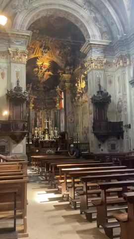 The video shows the interior of a church. The camera pans across rows of wooden pews. At the front of the church, there is an altar with religious symbols. Above the altar, there is a large fresco depicting religious figures. There are also ornate columns and wall paintings throughout the church. One person is visible sitting in the pews. The lighting suggests it is daytime, with sunlight streaming through the windows.