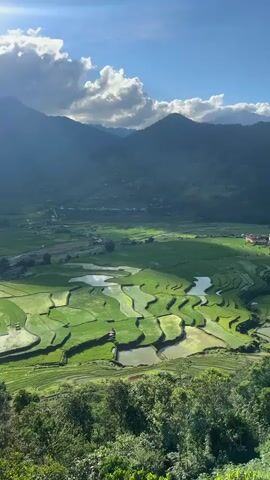 A high-angle, vertical shot captures a vast, terraced landscape with lush green rice paddies reflecting sunlight. In the distance, a narrow river winds through a valley, flanked by small buildings and settlements. Towering mountains under a partly cloudy, bright sky form the backdrop. The camera slowly zooms in on the paddies and the valley floor.