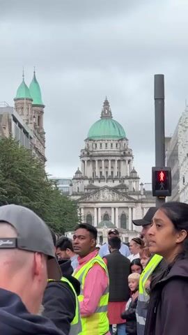 A crowd of people stand on a street, with a large, ornate building with a green dome in the background. Many people in the foreground are wearing high-visibility vests. A traffic light for pedestrians shows a red "don't walk" signal. The sky is overcast.