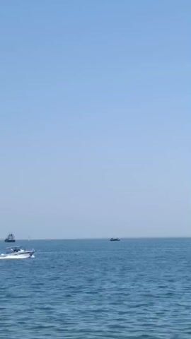 The video shows a wide view of the sea under a clear blue sky. Several boats are visible. In the foreground, a small boat with an outboard motor is moving to the right, with a person in it. In the middle distance, two larger ships are sailing. Further in the distance, a coastline with trees is visible. The water is calm with gentle waves. The overall impression is of a peaceful day at sea.