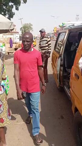 Here's a concise description of the video:

The video shows a street scene in what appears to be a city in Nigeria.  A woman in a red dress stands near a yellow and white minivan with its door open.  A man in a striped shirt and cap stands next to the van. Another person is partially visible near the camera. The video then pans to show the street, with other yellow auto rickshaws and cars. Buildings line the street on both sides.  The weather is sunny.