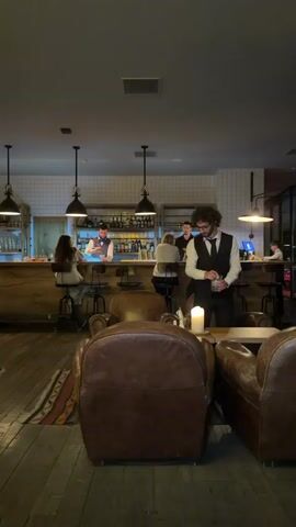 The video shows the interior of a dimly lit bar or lounge. In the foreground, two plush leather armchairs are visible, with a small, patterned rug on the wooden floor between them. A lit candle sits on a table in front of one of the armchairs. In the background, a long wooden bar with a white tiled backsplash stretches across the scene. Several bottles of alcohol are displayed on shelves behind the bar. Three bartenders are working behind the counter. One bartender, wearing a vest and bow tie, is visible on the far right. Another bartender, also in a vest and bow tie, is in the center, appearing to pour a drink. A third bartender is partially visible on the left. Two patrons are seated at the bar, facing the bartenders. One patron is on the left, and another is further to the right. The overall atmosphere appears to be sophisticated and relaxed.