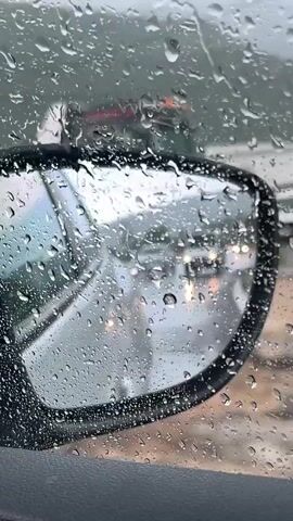 The media file shows a close-up view through a car window on a rainy day. Raindrops heavily coat the glass, distorting the view of the side-view mirror and the road beyond. The mirror reflects a street scene with other vehicles and lights, their details blurred by the water. The weather is clearly rainy and overcast, contributing to a muted color palette. The setting appears to be a street or road, possibly in a town or city, with buildings visible in the background through the rain-streaked window. The overall mood is one of enclosure and perhaps a touch of melancholy due to the inclement weather. There is no visible text in the media file. The location context provided is Podlastva (Poljice), Montenegro.