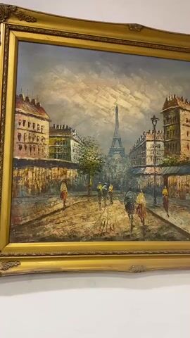 The video displays a framed oil painting of a Parisian street scene with the Eiffel Tower in the background. Several figures, possibly pedestrians and cyclists, are depicted walking or riding on the cobblestone street. The buildings on either side of the street have a distinct architectural style. The sky is rendered with dramatic clouds, and the overall impression is one of a slightly overcast day. The painting is framed with an ornate gold-colored frame. The camera is held steady, and the painting is the sole focus of the video.