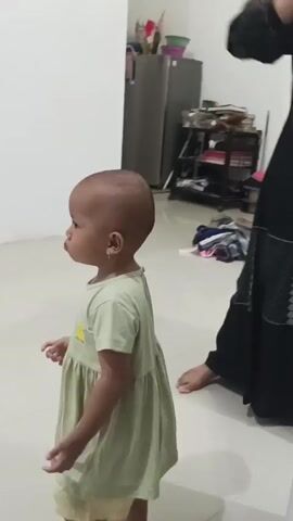 A toddler in a pale green dress walks to the left of the frame. The toddler is barefoot and has short hair. Behind the toddler, a person wearing a light brown hijab and an orange and brown top is visible, reaching up towards their head. The background shows a room with tiled flooring and white walls. To the right, a refrigerator and a shelf unit are visible, with various items on the shelves. To the left, a portion of a wooden door frame and what appears to be a bed with red and white striped bedding can be seen. A yellow water bottle with a pink lid is on the floor in the lower right corner of the frame.