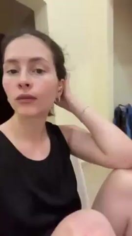The video shows a woman sitting indoors. She is fair-skinned with dark hair pulled back and wearing a black tank top. She has jewelry in her ear and a bracelet on her left wrist. Her arms and legs are bare. She appears to be sitting on a chair or low surface. The background includes beige walls and a partly visible clothes drying rack with clothes on it. A door and possibly a backpack are also visible. The camera is held at a low angle and pointed up at the woman, who looks directly at the camera for a few seconds. She briefly touches her hair.