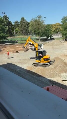 The video shows a construction site outside a modern, light green, multi-story building. In the foreground, a dark gray ledge is visible, suggesting the footage is being filmed from a window or balcony. The construction area is mostly dirt and gravel, with piles of material scattered throughout. 

On the left side of the construction site, a yellow excavator is actively digging, with its arm raised and bucket full of dirt. A person wearing a bright orange vest and a hard hat stands near the excavator, observing the work. Further into the site, another construction vehicle, a yellow loader, is parked near a white car. A small white car is also parked in the lot. 

The background features a clear blue sky and lush green trees. In the distance, there appear to be sports fields or courts with lights and a fence. The building itself has numerous windows and is labeled with text that appears to be in a foreign language. Signs on the building indicate it is a school. The overall impression is a sunny day at a school undergoing construction.