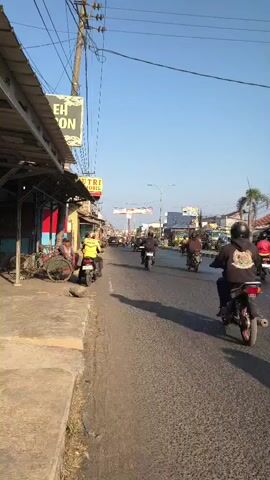 A street scene with a clear blue sky. In the foreground, a sidewalk runs along the left side of the image, with a covered market stall extending over it. A bicycle is parked against the stall. On the road, several motorcycles are moving away from the camera. A person in a yellow jacket is riding a motorcycle on the left side of the road, close to the sidewalk. Other motorcycles are further down the road, and some appear to be carrying passengers. The road is paved and shows signs of wear. Utility poles with wires are visible against the sky. In the distance, buildings and more vehicles are present, including a large truck on the right side of the road. The overall impression is of a busy street in what appears to be a town or city.