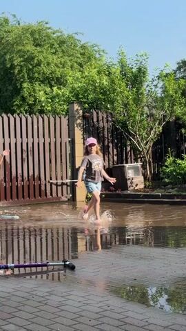 A young girl with blonde curly hair, wearing a pink baseball cap, a gray t-shirt, and a denim skirt, runs through shallow puddles on a brick pathway. She jumps with her arms raised. A scooter with a purple handlebar lies on the pathway in the foreground. A wooden fence and lush green trees form the background, with a gate visible on the fence. The sky is clear and blue.
