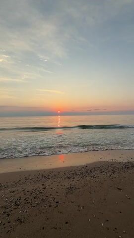 The video shows a sunset over the ocean. The sun is a small, bright orange ball positioned slightly to the left of the center of the frame, just above the horizon. Its reflection creates a shimmering path of light on the calm water. Small waves gently lap onto a sandy beach in the foreground, which is covered in shells and pebbles. The sky is a gradient of colors, transitioning from a pale blue at the top to soft pinks and oranges closer to the horizon. There are a few wispy clouds scattered across the sky. The overall scene is serene and peaceful.