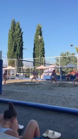 This video captures a beach volleyball game being played on a sand court. The subjects are several women actively participating in the sport, dressed in athletic wear, likely bikinis or sports bras and shorts. Two players are on one side of the net, and at least two are on the other. The scene is set on a sunny day, as indicated by the bright light and clear blue sky. Two tall, slender cypress trees stand prominently behind the court, suggesting a Mediterranean or similar climate. In the background, there are white umbrellas providing shade, a fence surrounding the court, and distant mountains, consistent with the provided location context of Gošići, Montenegro. A few spectators are visible sitting on a bench in the foreground, partially obscuring the lower portion of the court. The mood appears to be energetic and focused, typical of a sporting event. Visible text includes "ELITE" on a banner on the left side of the court and a blue "B" logo on a structure behind the net. The time of day is likely afternoon, given the angle of the sun casting shadows and the bright illumination.