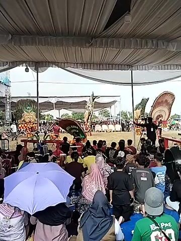This is a video recording of a cultural performance taking place outdoors, under a large white tent. The event appears to be a traditional Indonesian dance or festival, possibly a "barong" dance given the elaborate costumes and masks.

**Subjects:**
The primary subjects are performers in vibrant, elaborate costumes. One performer in the center wears a large, winged headdress that is red and green, with the words "NURUL HUDA SRAGEN" visible on a green banner attached to it. To the left and right of the stage are other performers or large costume figures. The left figure is a lion-like creature with a headdress of green foliage. The right figure is partially obscured but appears to be similar in style. The performers are engaged in energetic movements.

In the foreground, an audience is seated, with many people holding umbrellas to shield themselves from the sun or potential rain. A light purple umbrella is prominently displayed in the lower left of the frame. The audience members are diverse in age and are watching the performance.

**Activity or Event:**
The video captures a live performance, likely a traditional dance or theatrical presentation. The performers are moving dynamically, and the elaborate costumes suggest a significant cultural event. The audience's rapt attention indicates they are actively engaged in the performance.

**Notable Details:**
The time of day appears to be daytime, with bright lighting suggesting it could be mid-morning or afternoon. The presence of umbrellas might indicate sunny or possibly unpredictable weather. The setting is an open field, possibly a village square or community space, given the rural feel and the presence of tents set up for the event. The performance space is marked by a low wooden railing.

**Visible Text:**
The most prominent visible text is "NURUL HUDA SRAGEN" on the central performer's headdress.