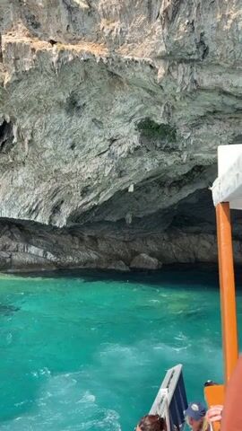 The video shows a boat sailing along a rocky coastline with turquoise water. The camera is positioned on the boat, looking towards a large cave opening in the cliff face. The cave appears dark and mysterious, with some stalactites hanging from the ceiling. The water is clear and a vibrant shade of blue-green, with small waves breaking against the shore. The sun is shining, and the sky is a clear blue. In the foreground, the edge of the boat is visible, with a few people on board. The boat is moving slowly, and the camera pans across the scene, capturing the natural beauty of the coastline and the cave. The video appears to be a travelogue or a scenic tour, showcasing a beautiful natural landscape.