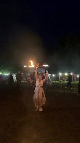 A woman in a light-colored dress and boots is performing with a flaming hula hoop in a dirt clearing at night. She is spinning the hula hoop around her waist and arms. There are spectators in the background, some standing and some sitting. There are tents and trees in the background. There is a fire in the foreground. The scene appears to be at a festival or event.