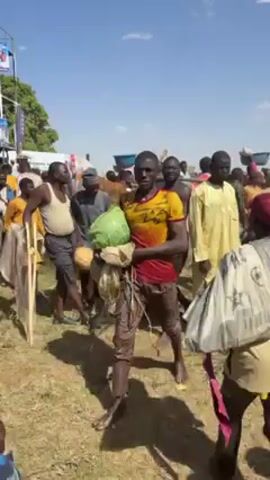The video shows a crowd of people, mostly men, walking in an outdoor area on a sunny day. Many of them are covered in mud. Some are carrying items such as a watermelon, a net, and a red canister. There is a tall, multi-level structure in the background, possibly a viewing platform, with advertising on it. The ground appears to be grassy or dusty. The people are moving in the same general direction.