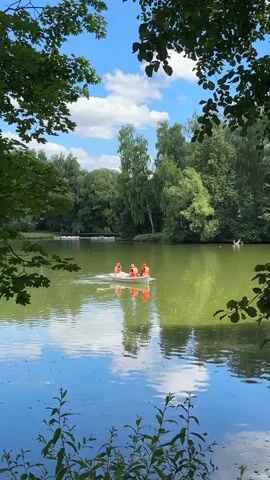 A small, white rowboat with three people wearing orange life vests is on a green, reflective lake surrounded by lush green trees under a blue sky with white clouds. The people are rowing the boat. The scene is framed by dark green leaves in the foreground.