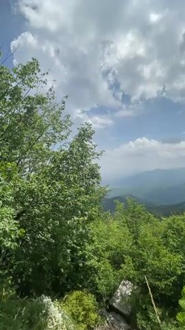 A drone perspective shot pans across a vibrant, lush green landscape of rolling hills and mountains. The foreground is dominated by dense foliage and trees. In the background, layers of mountains recede into the distance, becoming softer and more muted in color due to atmospheric perspective. The sky is a dynamic mix of bright blue patches and large, fluffy white clouds, suggesting fair weather with some cloud cover. The overall impression is one of natural beauty and expansive vistas.