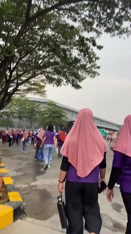 A group of women wearing purple shirts and pink headscarves are walking in a line on a paved path. One woman in the foreground is carrying a black water bottle. They are walking away from the camera, towards a large industrial building in the background. The path is lined with trees and a yellow and black striped curb. The sky is overcast.