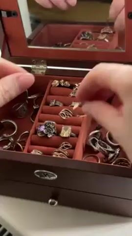 A pair of hands are shown reaching into a brown jewelry box that has a mirror attached to the inside of the lid. The jewelry box is lined with red velvet and filled with rings. The hands pick up a silver ring with a light purple stone, place it on a finger, and then remove the ring and place it back into its spot in the box. The jewelry box is sitting on a white surface.