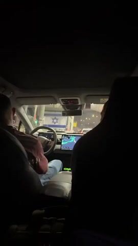 The video, taken from inside a car at night, shows the interior of a car with two people. The person in the driver's seat, facing forward, is wearing a red shirt and blue jeans. The other person is in the passenger seat, facing the driver. The car has a large touchscreen display in the center console which shows a navigation map. The Israeli flag is prominently displayed hanging from the rearview mirror. The background outside the car is dark and appears to be a road with some lights and structures visible. The video is short and appears to be a casual recording.
