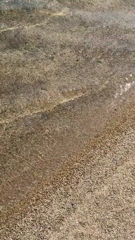 A video shows the edge of a body of water lapping against a sandy and pebbly shore. Footprints are visible in the sand near the water's edge. The water is clear and reflects sunlight, creating shimmering patterns. The scene is captured from a slightly elevated, close-up perspective, focusing on the interaction between the water and the land.
