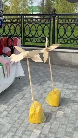 The video shows two wooden bird-shaped pinwheels standing on a brick-paved area. They are attached to wooden poles and stand in yellow plastic bags, likely filled with weight for stability. In the background, there's a black wrought iron fence and a glimpse of a green lawn. To the left, partially visible, is a vendor's table with some merchandise including hats and scarves on mannequins. The wind is gently turning the pinwheels. The scene appears to be outdoors on a sunny day.