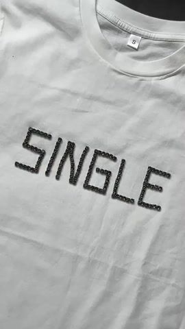 The video displays a close-up, overhead shot of a white t-shirt with the word "SINGLE" spelled out in dark, sparkly rhinestones. The t-shirt is laid flat on a surface, and the camera slowly zooms in on the word. The background is a plain white surface, likely a table or the floor. There are no people visible in the video. The lighting is bright, highlighting the texture of the t-shirt and the shimmer of the rhinestones. The video is short, lasting only a few seconds, and the movement is primarily the camera zooming in and slight panning.