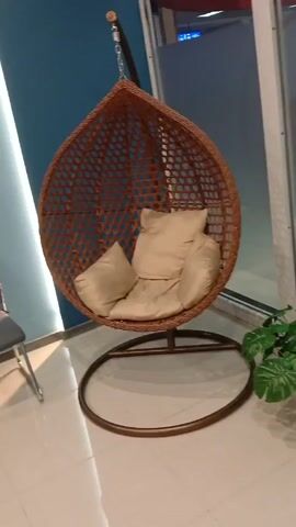 The video shows a hanging egg chair inside a room. The chair is made of brown wicker and has beige cushions inside. The chair is suspended from a metal chain attached to a point above the frame. The base of the chair is a circular metal stand. To the left of the hanging chair is a grey chair with metal legs. To the right of the hanging chair is a potted plant. The background includes a blue wall on the left and a window or glass wall on the right. The floor is light-colored tile. There is a slight movement of the camera but overall the scene is still.