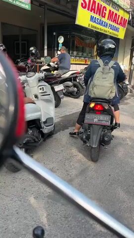 A low-angle shot captures a street scene where several motorcycles are parked. In the foreground, the side mirror of a motorcycle is visible. In the middle ground, a white scooter is parked to the left, and a person wearing a helmet and a backpack is seated on a black motorcycle to the right, facing away from the camera. The motorcycle has a license plate that reads "W 2577 SH". Another person wearing a cap is seated on a white motorcycle behind the black motorcycle. In the background, a yellow sign with red lettering that says "SIANG MALAM MASAKAN PADANG" is visible. The environment appears to be a street in a commercial area, with buildings lining the street. The lighting suggests it is daytime.