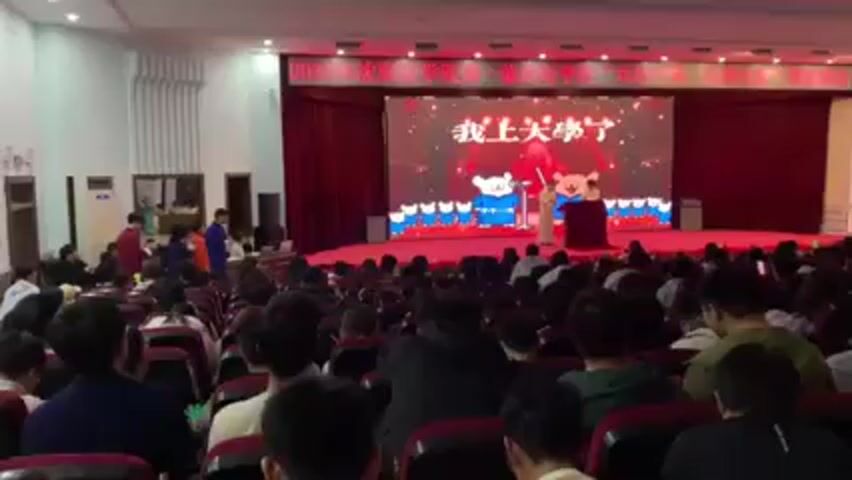 An auditorium is filled with seated people, likely students, facing a stage with two performers. A large screen behind the stage displays a cartoonish image with Chinese text. The performers, one at a podium, are dressed in traditional robes. A few individuals stand or walk near the entrance of the auditorium. The audience appears attentive, with some holding up props.