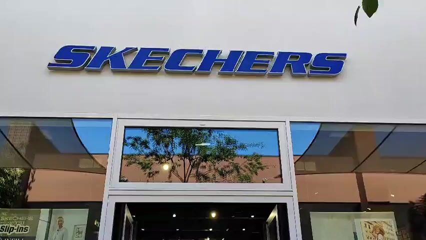 The video shows the exterior of a Skechers shoe store on a sunny day. The store has a prominent blue "SKECHERS" sign above a large glass entrance. Through the glass, a variety of shoes are displayed on shelves inside the store. Several people are seen walking in and out of the store, and some are browsing the merchandise. The windows on either side of the entrance also display advertisements for Skechers products. The overall environment appears to be a commercial street with other shops and businesses visible in the background.