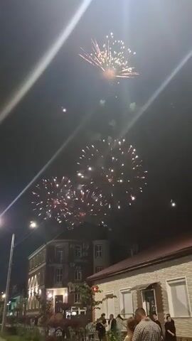 The video shows a night scene with fireworks exploding in the sky. Below the fireworks, there are buildings and a street with people. In the foreground, a couple is standing near a building. There are streetlights and some trees visible. The fireworks are colorful, with bursts of white, pink, and red light. The people appear to be watching the fireworks display. The environment seems to be a town or a city street.