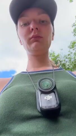 The video starts with a close-up of a young woman wearing a green tank top with navy and white trim. She has a black baseball cap on and a DJI camera hanging from her neck. The camera zooms in on the camera, and then back to her face. The woman makes a face and then smirks. In the background, a man walks away from the camera wearing a white shirt, maroon pants, and carrying a red sweater. The camera then shows a wide shot of a valley with lush green foliage and mountains in the background, with a blue sky and fluffy white clouds overhead.