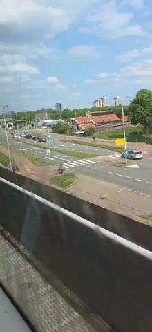 The video captures a roadside view from what appears to be a moving vehicle. A road with several lanes runs parallel to the vehicle, with cars traveling in both directions. A person on a bicycle is seen crossing the road at a pedestrian crossing. The background features buildings, trees, and a partly cloudy sky. The vehicle's interior is visible in the foreground, partially obscuring the view.