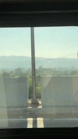 The video shows a view through a window, possibly from a vehicle. In the foreground, there is a concrete barrier with some torn fabric hanging from it. The fabric appears to be white and translucent. Below the barrier, a road surface is visible with some debris. The background shows a hazy landscape with buildings, trees, and mountains in the distance. The sky is clear and blue. The overall impression is of a distant view of a town or city in a dry, possibly desert, environment. The time stamp indicates a duration of approximately four seconds.