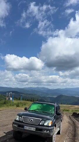 A black SUV is parked on a dirt road with a mountain range in the background under a cloudy blue sky. The video then pans to show three people, two men and a woman, standing on a hillside with the mountains behind them. Next, the camera focuses on two pine trees on a grassy hill with the mountain range in the distance. The video then pans to show a ski lift structure on a mountainside with trees and mountains in the background. Finally, a woman wearing a cowboy hat, sunglasses, a black tank top, and light-colored pants walks across a grassy hill with mountains and a cloudy blue sky in the background.