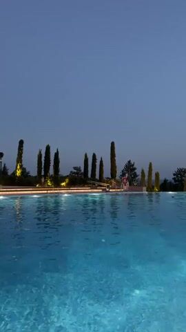 The video shows a large, illuminated swimming pool with clear blue water. Tall, slender trees line the edge of the pool, with artificial lights illuminating them and the surrounding area. A multi-story building with balconies and lit windows is visible in the background. Several people are seen swimming or wading in the pool, and some are relaxing by the poolside. The sky is a deep twilight blue, suggesting it is either early evening or late dusk. The overall atmosphere appears serene and luxurious.