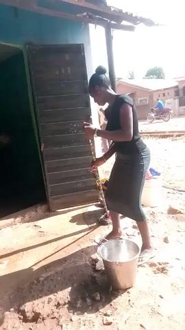 A woman is mopping the area outside of a building. The building has a turquoise wall and a dark, slatted door that is partially open. The woman is wearing a black top, a gray skirt, and white sandals. Her hair is pulled back in a bun. She is holding a mop with a green handle and a gray mop head. Next to her is a silver bucket and a dark trash can. The ground is dirt and there are some weeds growing. In the background, there are other buildings and trees.  The weather looks sunny. The woman dips the mop into the bucket, then wrings it out over the bucket, and then straightens up and looks directly at the camera.