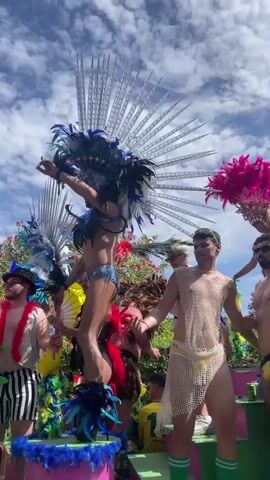The video shows a parade float decorated with vibrant colors and feathers. Several individuals are present on the float, some wearing elaborate costumes with large feather headdresses and sparkling attire. One person is dressed in a mesh top and yellow briefs, another in yellow briefs and a shirtless torso, and another in white pants, a sparkly vest, and a white hat, adorned with green and pink feathers. A woman with large butterfly-like wings and a feathered boa stands near a DJ setup. The float has banners that read "Queenz" and "Outlet Sitges." The sky above is partly cloudy, and lush green foliage is visible in the background. The people on the float appear to be dancing and interacting, likely part of a celebration or festival.