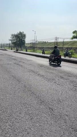The video shows a wide shot of a paved road on a sunny day. The road has many potholes. A person is riding a motorcycle in the middle lane. There are other motorcycles in the distance. On the right side of the road, there is a grassy embankment with some trees and power lines. The sky is clear and blue.