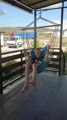 A young boy with blond hair, wearing a black t-shirt with a graphic design and patterned shorts, is sitting in a blue hammock chair. The chair is suspended from a metal frame under a covered porch with a wooden floor and railing. The boy is swinging gently back and forth, looking towards the camera with a smile. In the background, there is a white truck with a red cab, a blue trailer, and houses on a hill under a bright, cloudy sky. The overall setting appears to be a residential outdoor area in a rural or semi-rural location.