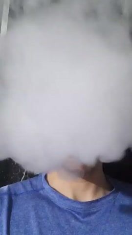 A person wearing a blue t-shirt is obscured by a large cloud of white vapor. The person's face is not visible. The background is dark and indistinct.