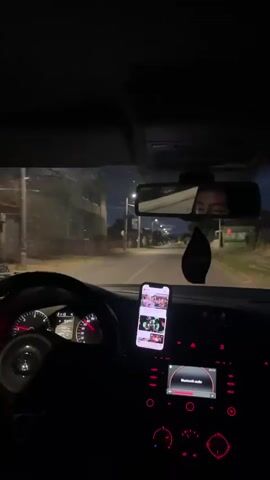 A person is driving a car at night, likely in a residential area, listening to music through the car's Bluetooth audio system. The dashboard is illuminated, with the speedometer showing the car is in motion. A smartphone is mounted on the dashboard, displaying content, possibly social media or a playlist. The reflection in the rearview mirror shows a person's face, suggesting they are the driver. The street is illuminated by streetlights, and there are houses visible along the roadside. The lighting is generally dim, creating a nighttime ambiance.