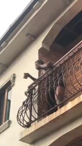 A person is standing on a balcony. The person is dark-skinned and wearing dark clothing. They are holding a brown mug. The balcony has an ornate black metal railing and a brick wall. The building is light-colored with a window to the left of the balcony. The sky is overcast. The person raises the mug to their lips and drinks. They then lean over the railing and look down.