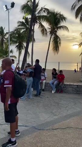 The scene captures a group of people gathering near a basketball court set up outdoors, likely for a game or event. Several palm trees frame the background, suggesting a beachside or tropical location. The sky is overcast, indicating either early morning or late afternoon light.

In the foreground, several individuals stand observing the court.  Two men in the left corner are particularly noticeable; one wears a red-and-orange patterned shirt and a crossbody bag, while the other wears a maroon shirt with a backpack. To their right, a small crowd is loosely gathered, some seated on a low wall. Further on, two women stand side by side, seemingly watching the game. One is dressed in jeans and a white shirt, and the other wears jeans and a t-shirt with an image and writing.

The basketball court is the focal point, marked with black and gray sections and featuring a banner with text partially visible. Several people stand and mill about the court.  Three women in matching maroon basketball uniforms walk onto the court. In the foreground, a man wearing blue shorts with yellow lightning patterns stands watching the court.

Behind the basketball court, buildings rise in the distance, including a hotel. This mix of urban and natural elements confirms the location as a coastal area or city park.