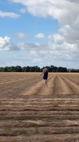 A young woman with blonde hair, wearing a dark blue dress, runs with her arms outstretched in a field. The field is covered with rows of dried, golden-brown grass or hay. In the distance, a line of trees forms a horizon against a bright blue sky with scattered white clouds. The woman is running away from the camera, down the center of a strip between two rows of crops. The camera is stationary, and the video is a continuous shot.