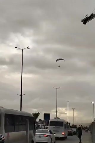 The video shows a paraglider flying low over a busy road on a cloudy day. The paraglider is silhouetted against the gray sky and appears to be descending. There are cars, vans, and pedestrians on the road below. Streetlights line the road and a construction crane is visible in the background. The camera follows the paraglider as it moves and gets closer to the light poles. People on the ground seem to be watching the paraglider.