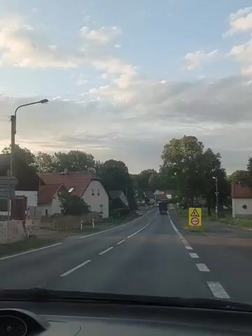 The video shows a view from the inside of a car, looking forward through the windshield. The car is on a road in a rural or suburban area, with houses and trees lining the sides. There are traffic signs visible, including a yellow triangular sign with an image of children and a circular sign with a red border and a diagonal red line. In the distance, there is a police car with flashing blue lights, and other vehicles. The sky is blue with some clouds. The video is shaky, suggesting the car is moving.