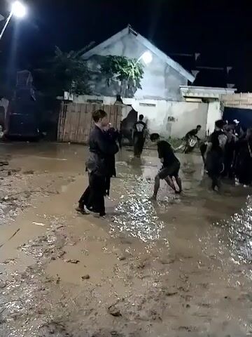 At night, an outdoor scene unfolds in a muddy, unpaved area, likely after rain or intentional watering. Several people, mostly men, are gathered, illuminated by artificial lights. Many are dressed in black attire, resembling martial arts uniforms, while one individual in the foreground is wearing a vibrant red sarong-like garment.

The central activity involves the person in the red garment moving actively in the mud, seemingly performing a dance or martial arts routine, while others observe or participate. Some individuals are seen holding what appear to be whips or ropes. In the background, there is a blue tarp structure, a flag (possibly the Indonesian flag) on a pole, and residential buildings. The ground is visibly wet and slushy, with puddles reflecting the lights. The atmosphere suggests a traditional cultural performance or martial arts demonstration, possibly Pencak Silat, given the attire and movements. Traditional Indonesian music accompanies the scene.